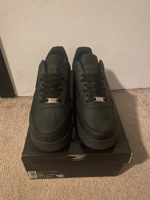 Svarta Nike Air Force 1 - EU 43 - Klassiska helsvarta Nike Air Force 1 sneakers i lågt utförande. Skorna har ovandel i läder, perforerade detaljer på tån och den ikoniska Swoosh-loggan på sidan. Tjock sula och metallbricka på snörningen ger en clean och tidlös look. Använda endast 3 gånger sparsamt. Skolåda tillkommer INTE