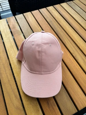 Ljusrosa klassisk keps - Snygg ljusrosa keps i klassisk modell med böjd skärm och justerbar rem bak. Tillverkad i mjukt bomullstyg och har ventilationshål på ovansidan för extra komfort. Perfekt accessoar för en avslappnad och trendig look.