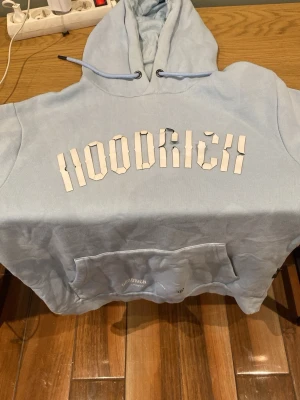 Ljusblå HOODRICH hoodie med huva - Snygg ljusblå hoodie från HOODRICH med stor logga i vitt på bröstet. Klassisk känguruficka framtill och justerbar huva med dragsko. Perfekt för en chill och avslappnad stil. Tillverkad i mjukt material för extra komfort.