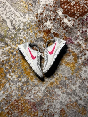 Nike vita sneakers med rosa swoosh - Nike sneakers i vitt meshmaterial med gråa detaljer och en tydlig rosa swoosh på sidan. Skorna har vita snören, grå hälparti och en flexibel vit sula med svarta partier undertill. Perfekta för en sportig och avslappnad stil.