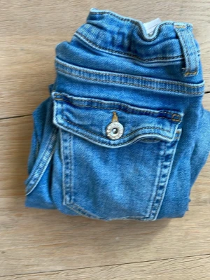 Blå bootcut jeans med fickdetalj - Säljer ett par klassiska blå bootcut jeans i denim med snygg ficka bak och knappdetalj. Jeansen har normalhög midja och utsvängda ben, perfekta för en avslappnad och trendig look. Passar dig som gillar retrovibbar och tidlös stil. Priset går också att diskuteras❤️🫶🙌💕
