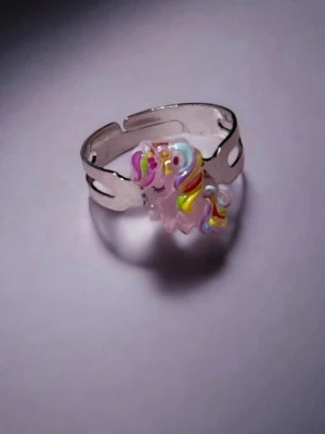 Söt ring med enhörning och regnbågsfärger - fler färger finns - Unik ring i silverfärgad metall med en liten rosa enhörning som dekoration. Enhörningen har färgglad man i regnbågens färger och blundande ögon, vilket ger en lekfull och gullig look. Perfekt för dig som älskar fantasifulla accessoarer och vill sticka ut. Går att få ringen i guld, föer färger på enhörning finns också. Ringen går att göra mindre och större själv. 