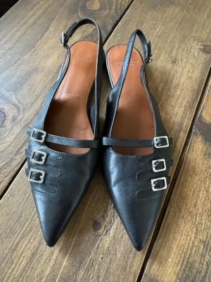 Svarta ballerina Vagabond - Snygga svarta ballerina från Vagabond, Hermine, med spetsig tå och tre dekorativa silverfärgade spännen på varje sko. Skorna har öppen häl med justerbar rem och är tillverkade i slätt läder. Perfekta för dig som gillar en edgy men stilren look. Knappt använda. Nypris 1100kr