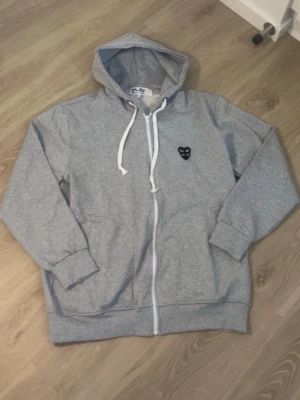 Grå hoodie Comme des Garçons PLAY - Säljer en grå zip-hoodie från Comme des Garçons PLAY i storlek XL. Hoodien har vit dragkedja, vita snören och den ikoniska svarta hjärt-loggan på bröstet. Tillverkad i mjuk bomullsblandning och har huva samt ribbade muddar vid ärmar och nederkant.kom med frågor och funderingar🫶🏻😙