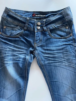 Blå jeans från Redseventy Jeans low waist 2000s - Säljer ett par klassiska blå jeans från Redseventy Jeans med dubbla silverknappar och femficksmodell. Jeansen har snygga slitningar och kontrastsömmar som ger en cool look. Perfekta till vardags och matchas lätt med olika stilar.