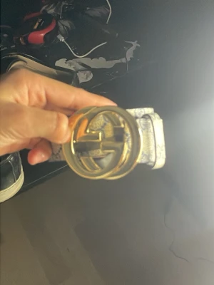 Gucci bälte med GG-mönster och guldspänne - Säljer ett snyggt Gucci bälte med klassiskt GG-mönster i blått på en ljus bakgrund. Bältet har ett stort, rundat spänne i guld med GG-logga. Perfekt för att lyfta din outfit med en lyxig touch. Materialet är skinn.