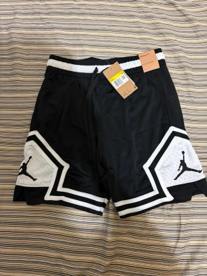 Jordan shorts - Snygga svarta Jordan shorts med vita kontrastdetaljer och klassisk Jumpman-logga på båda benen. Shortsen har elastisk midja med snörning och är perfekta för sportiga outfits. Tillverkade i ett lätt och ventilerande material.