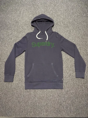 Mörkblå hoodie från Superdry - Säljer en mörkblå hoodie från Superdry med grön logga på bröstet. Hoodien är i storlek S