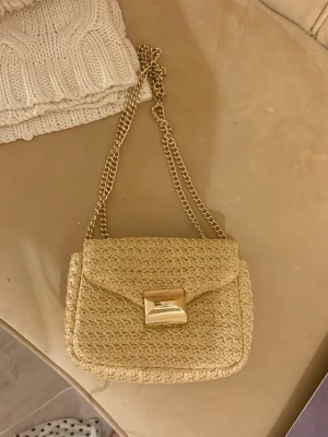Beige flätad handväska från H&M - Snygg beige handväska från H&M i flätad strådesign med guldfärgad kedjerem och ett stilrent guldfärgat spänne framtill. Perfekt accessoar för dig som gillar trendiga detaljer och naturliga material. Väskan är verkligen i helt nytt skick!🙌🙌🙌