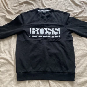 Hugo Boss Sweatshirt - Svart sweatshirt från Hugo Boss med stor vit logga över bröstet. Klassisk rund halsringning och långa ärmar. Tröjan är i mjuk bomull och har en relaxed passform som passar perfekt till jeans eller joggers.