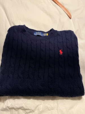 Mörkblå kabelstickad tröja Polo Ralph Lauren - Kabelstickad mörkblå tröja från Polo Ralph Lauren med rund halsringning och röd broderad logotyp på bröstet. Tröjan har klassisk passform och ribbstickade muddar vid hals, ärmslut och nederkant. 