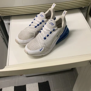 Nike Air Max 270 vita/blå sneakers - Nike Air Max 270 sneakers i vitt med blå detaljer och stor blå air-bubbla i hälen. Ovandel i mesh för bra andning, snörning och tydlig Air 270-logga. Snygg sportig look och bekväm passform, perfekt för dig som gillar streetstyle och sneakers med attityd. Slitet på höger sko vid lilltån 