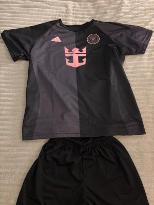 Svart och rosa Messi fotbollströja - Säljer en svart och rosa fotbollströja från Adidas med Messi och nummer 10 på ryggen. Tröjan har korta ärmar, rund hals och är gjord i ett lätt, andningsbart material. Snygg design med klubbmärke och logga på bröstet. Matchande svarta shorts ingår.