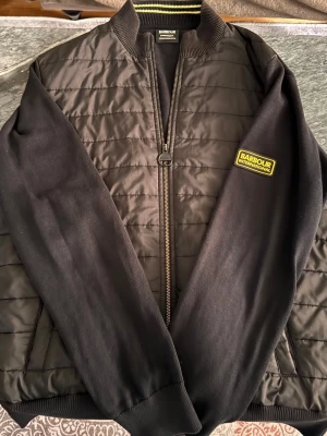  Barbour jacka  - Snygg svart bomberjacka från Barbour International med quiltad front, ribbade muddar och krage med gul detalj. Jackan har dragkedja och en gul logopatch på bröstet. Materialet är en mix av vadderad polyester och bomull för extra komfort. Super skick, ny pris kostar den mer eller mindre 2000kr men jag säljer den för 750