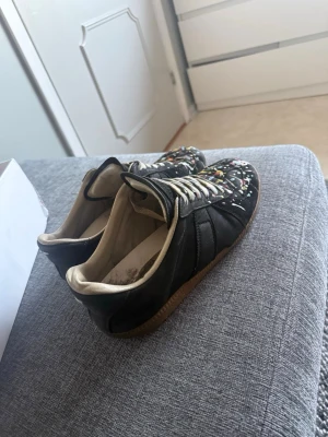 Svarta sneakers med färgstänk - Unika svarta sneakers med färgstänk i vitt, gult, rött och blått över hela ovandelen. Skorna har beige detaljer och vita snören. Snygg kontrast mellan den mörka basen och de färgglada detaljerna. Perfekta för dig som vill sticka ut med din stil.