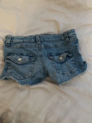 Blå jeansshorts med rå kant - Snygga blå korta jeansshorts med rå, fransig kant och klassiska fem fickor. Shortsen har bälteshällor, knapp- och dragkedjestängning samt dekorativa bakfickor med lock och knapp. Perfekta för varma dagar och en avslappnad stil.