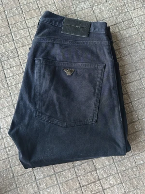 Mörkblå jeans/chinos från Emporio Armani - Snygga blåa jeans/chinos från Emporio Armani med klassisk femficksmodell och silverfärgade detaljer. Bakfickan har den ikoniska Armani-loggan i metall. Jeansen har normal midja och raka ben. Perfekta för dig som gillar stilrena och exklusiva plagg. Väldigt sparsamt använda. Köpta på Care of Carl.