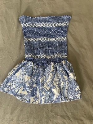 Blåvit axelbandslös kortklänning - Supersöt axelbandslös kortklänning i blått och vitt med smock upptill och volangig kjol. Klänningen har ett paisleyinspirerat mönster och är perfekt för varma dagar. Tillverkad i mjuk bomull som känns skön mot huden. Håller även sig på plats med gummibanden 