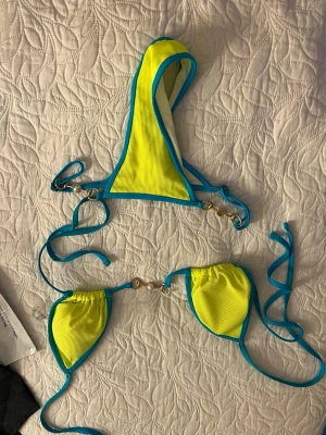 Gul och blå bikini med snörning - Trendig bikini i starkt gul färg med blå kanter och snörning. Toppen har trekantsform och dekorativa guldfärgade ringar mellan kuporna. Matchande underdel med samma färg och detaljer. Perfekt för stranden eller poolen!