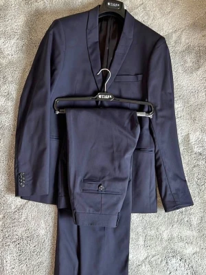 Mörkblå kostym från Tiger of Sweden - Snygg mörkblå kostym från Tiger of Sweden med klassisk tvåknäppt kavaj och matchande byxor.   Färg: Mörkblå (navy) Storlek: 48 Skick: Utmärkt – endast använd 1–2 gånger, som ny Tyg: Högkvalitativ ull från italienska väveriet Angelico Passform: Modern och stilren – sitter snyggt och bekvämt  ✅ Perfekt för bröllop, anställningsintervjuer, student, fest eller andra formella tillfällen. 🎯 Säljer då den bara hänger i garderoben.