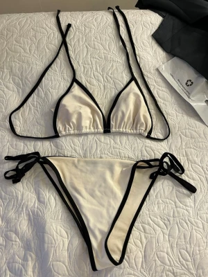 Vit bikini med svarta kanter - Snygg trekantsbikini i vitt med svarta kontrastkanter och knyt i både topp och trosa. Klassisk och stilren modell som passar perfekt till stranden eller poolen. Justerbara band för bästa passform.