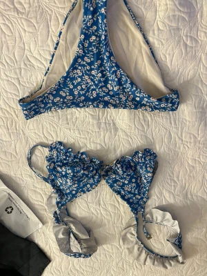 Blommig blå bikini med volang - Söt blå bikini med vitt blommönster och volangdetaljer på överdelen. Bikinin har knyt i sidorna och är perfekt för strandhäng. Materialet känns mjukt och bekvämt mot huden.