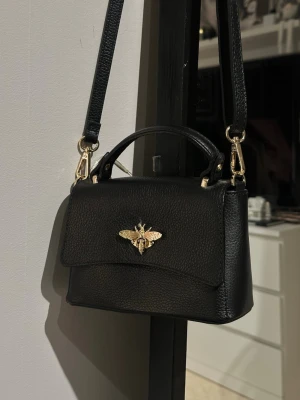 Svart handväska med gulddetalj - Snygg svart handväska i läderimitation med guldfärgad bi-detalj på framsidan. Väskan har både handtag och justerbar axelrem som går att ta av. Stilren och trendig modell som passar till många outfits.