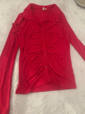 Röd långärmad topp - Trendig röd långärmad topp från Nelly med krage och knappar framtill. Toppen har snygg rynkning längs knappraden, mycket bra skick då den endast är använd en gång!