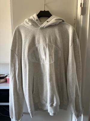 Ljusgrå hoodie från Risky - Ljusgrå hoodie från Risky med stor magficka och broderad logga på bröstet. Klassisk huva med dragsko och ribbade muddar vid ärmar och nederkant. Perfekt för en avslappnad och street-inspirerad stil. (Har original band till hoodien)