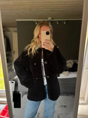 Svart teddyjacka med knappar - Mysig svart teddyjacka med  stora knappar. Jackan har en rak passform, två sidofickor och är gjord i fluffigt teddy-material 