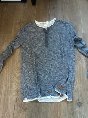 Blåmelerad långärmad tröja - Snygg blåmelerad långärmad henleytröja med knappar vid halsen och en bröstficka. Tröjan har vita detaljer vid krage och nederkant samt en liten läderpatch. Perfekt för en avslappnad stil.