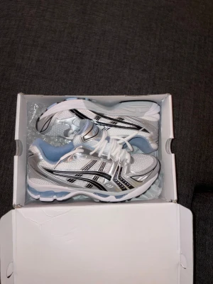 Asics Gel-Kayano 14 vit/svart sneakers - Snygga Asics Gel-Kayano 14 sneakers i vitt med svarta och silverfärgade detaljer. Ovandelen är i mesh och syntetmaterial, med blå insida och klassisk snörning. Sulan är vit med inslag av blått och silver. Perfekta för dig som gillar sportig och trendig stil.