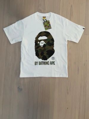 Vit BAPE t-shirt med camo-print - Säljer en vit t-shirt från A Bathing Ape med det ikoniska apansikte-trycket i camouflage på bröstet och texten 'BY BATHING APE' under. Klassisk passform och rund hals. Perfekt statement-plagg för streetwear-stilen.