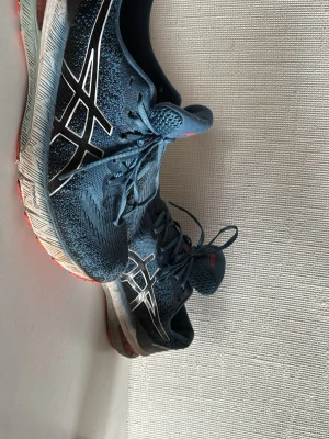 Blå Asics sneakers med vita detaljer - Snygga blå sneakers från Asics med vita och svarta detaljer på sidorna och röd sula. Skorna har klassisk snörning och mesh-överdel för bra andningsförmåga. Perfekta för dig som gillar sportig stil och vill ha bekväma skor till vardags.