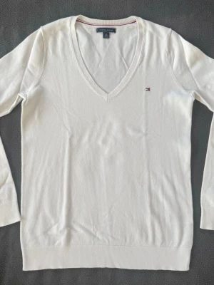Vit stickad tröja Tommy Hilfiger - Vit stickad tröja från Tommy Hilfiger med V-ringning och diskret logga broderad på bröstet. Tröjan har långa ärmar och ribbade muddar vid ärmslut och nederkant. Klassisk och stilren design i mjukt material.