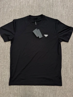 Svart t-shirt från Prada - Snygg svart t-shirt från Prada med klassisk passform och rund hals. Diskret logga på bröstet i form av en liten patch. Perfekt för en clean och stilren look.