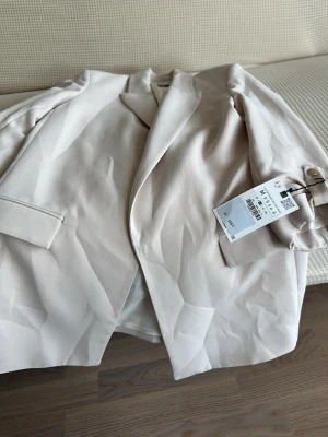 Beige kavaj från Zara, rak modell - Stilren beige kavaj från H&M i rak modell med klassiska slag och två fickor framtill. Kavajen är helfodrad och har en mjuk, slät yta. Perfekt för en clean och modern look.