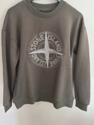 Stone Island sweatshirt med broderad logga - Säljer en snygg olivgrön sweatshirt från Stone Island med stor broderad logga i vitt på bröstet. Tröjan har rund halsringning och långa ärmar, perfekt för dig som gillar streetwear och vill ha något stilrent men ändå unikt.