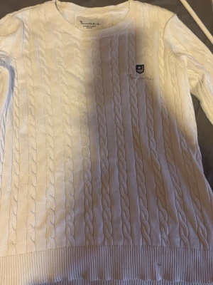 Vit kabelstickad tröja Bondelid - Kabelstickad vit tröja från Bondelid med rund halsringning och diskret logga på bröstet. Tröjan har ribbade muddar vid ärmslut och nederkant, perfekt för en klassisk och stilren look.