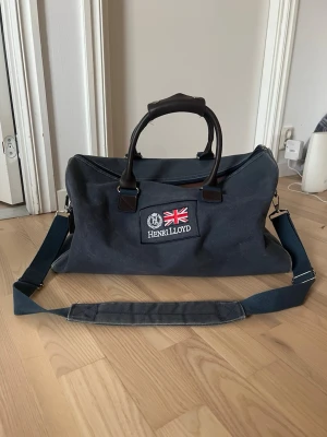Mörkblå weekendbag från Henri Lloyd - Snygg mörkblå weekendbag från Henri Lloyd med broderad logga och brittisk flagga på framsidan. Väskan har både handtag i läder och justerbar axelrem för extra komfort. Tillverkad i slitstark canvas, perfekt för resor och äventyr.