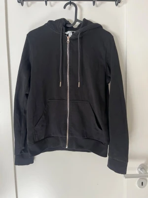Svart zip-hoodie med fickor - En svart hoodie med dragkedja framtill, två stora fickor och justerbar huva med snören. 