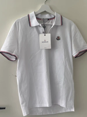Vit Moncler pikétröja med rödblå detaljer - Stilren vit moncler piké, perfekt för sommaren med väldigt bra kvalité, mycket bättre jämfört med den andra vita i min profil. Perfekt för dig som vill känna dig överfet med en riktig sommarvibe under sommaren.                               Se till att kontakta vid minsta lilla fundering