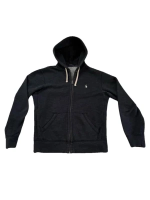 Polo Ralph Lauren Zip Sweater - Snygg mörkgrå hoodie med dragkedja från Polo Ralph Lauren. Klassisk design med vit broderad logga på bröstet, två fickor fram och vit dragsko i huvan. Mjuk insida och ribbade muddar för extra komfort. Perfekt för en chill och stilren look.