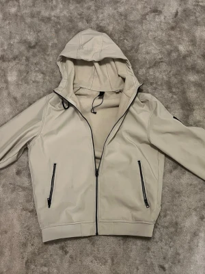 Beige vindjacka med huva - Stilren beige vindjacka med hel dragkedja framtill och två svarta dragkedjefickor. Jackan har en justerbar huva och ribbade muddar vid ärmslut och nederkant. Perfekt för blåsiga dagar och enkel att matcha med olika outfits.