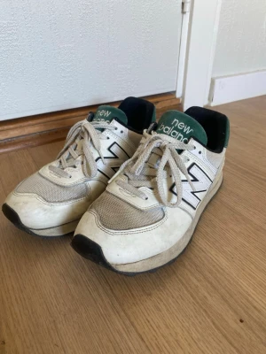Vita New Balance sneakers med gröna detaljer - Snygga vita New Balance sneakers med gröna detaljer och svart sula. Skorna har klassisk mesh och läder på ovandelen, stora N-loggor på sidorna och gröna tungor med vit text. Perfekta för en sportig och avslappnad stil.
