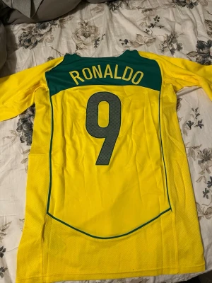 Brasilien Ronaldo 9 matchtröja Nike - Gul Brasilien fotbollströja med gröna detaljer, nummer 9 och namnet Ronaldo på ryggen. Tröjan har Nike-logga, Brasilien-emblem och är tillverkad i lätt, ventilerande material. Perfekt för dig som älskar fotboll och vill hylla en ikon.
