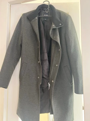 Mörkgrå kappa från Only & Sons - Stilren mörkgrå kappa från Only & Sons med klassisk krage och dragkedja samt knappar framtill. Kappan har två snedställda fickor och är fodrad för extra värme. Perfekt för kyliga dagar och ger en clean look till din outfit.