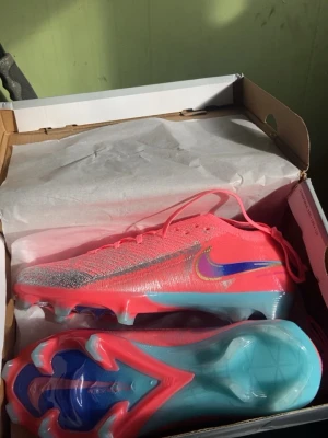 Nike Mercurial vapor 16 - Säljer ett par Nike Mercurial fotbollsskor i en riktigt snygg rosa färg med blå och silverdetaljer. Skorna har en låg profil, snörning och en yttersula i blått och rosa för bra grepp på planen. Ovandelen är i syntetmaterial med en glansig finish och Nike-logga i blått och gult.