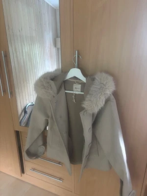 Beige kappa med fuskpäls från Only - Snygg beige kappa från gina med stor krage och fuskpäls runt halsen. Kappan har knäppning framtill och två fickor. Perfekt för kyliga dagar och ger en trendig look. Materialet är mjukt och behagligt.