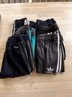 Adidas mjukisbyxor med ränder - Flera par klassiska Adidas mjukisbyxor i svart och mörkgrått med vita ränder längs sidorna. Byxorna har resår i midjan och är tillverkade i mjukt material, perfekt för en avslappnad stil. Några har även gröna detaljer och diskret logga på benet. Storlekarna är blandade från 9 till 11 år.